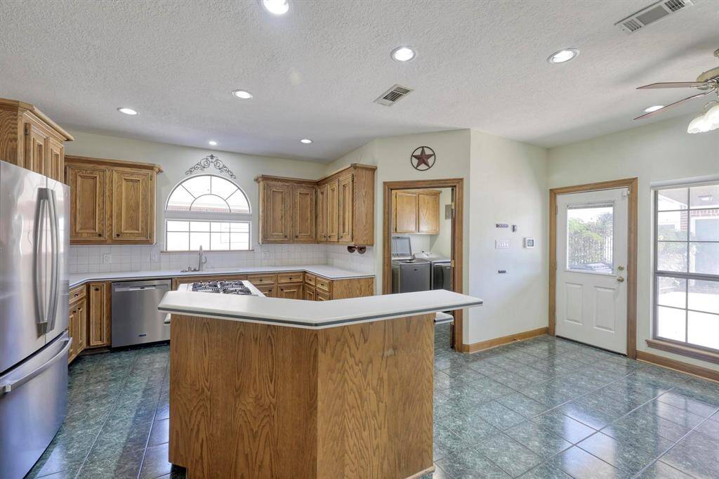 This kitchen features oak cabinetry, a central island with a cooktop, and stainless steel appliances. It offers ample natural light through large windows and a door leading outside. The space is accented by green tile flooring and recessed lighting, creating a bright and functional cooking area.
