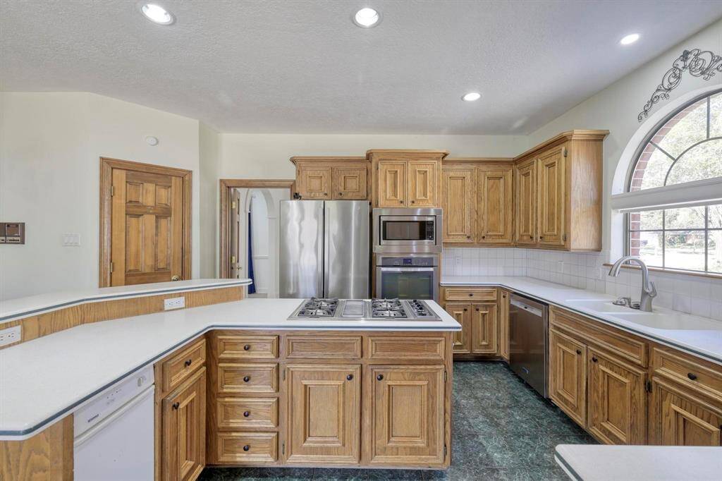 This kitchen features classic wooden cabinetry, modern stainless steel appliances, and a central island with a cooktop. It has ample counter space, a large arched window for natural light, and a clean, inviting design.