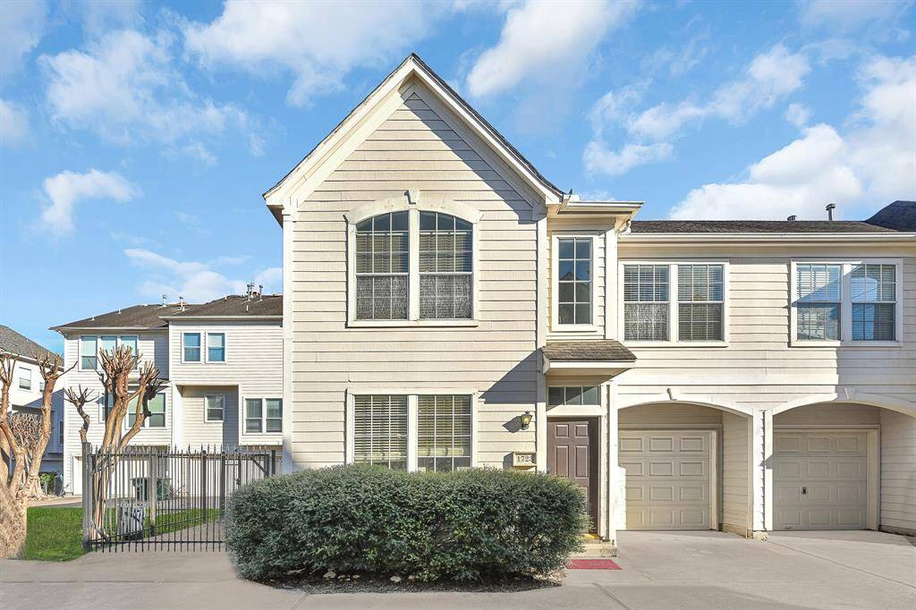 This charming two-story townhouse features a light-colored exterior with large windows and a gabled roof. It includes an attached garage and a small front yard with shrubbery, providing a cozy and inviting facade.