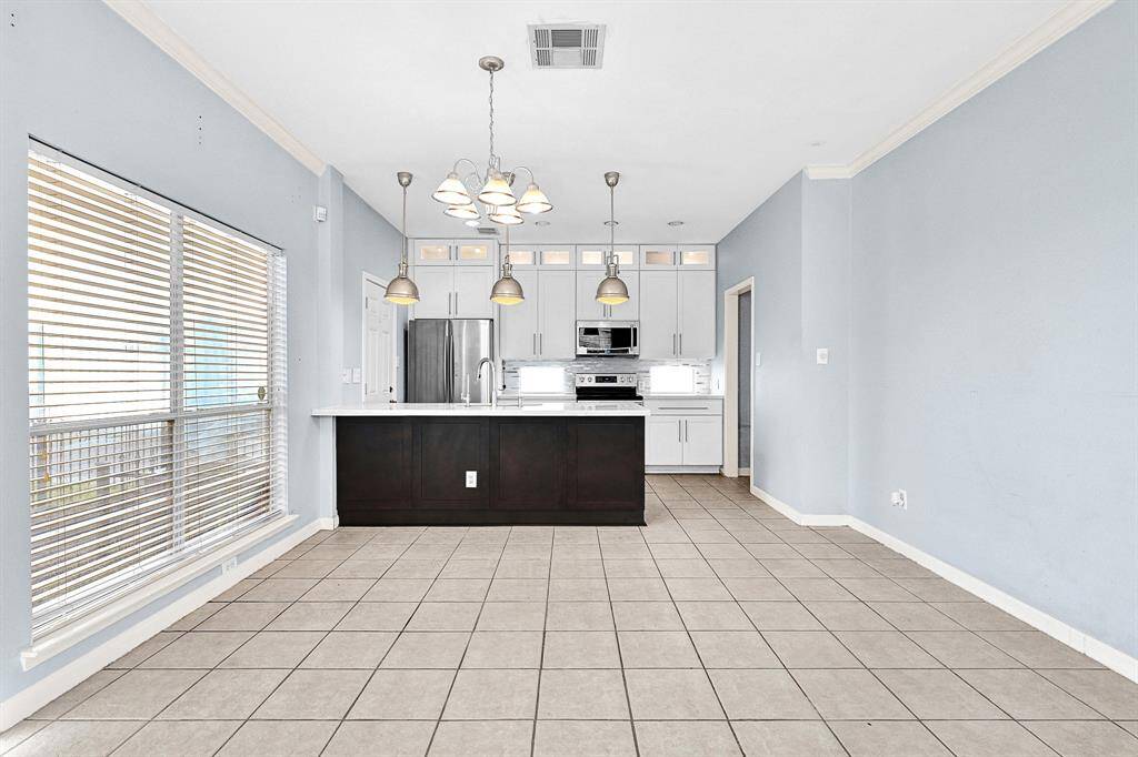 This is a bright, open-plan kitchen and dining area featuring light blue walls, large windows with blinds, and tiled flooring. The kitchen has modern white cabinetry, stainless steel appliances, and a central island with a dark base and pendant lighting above.
