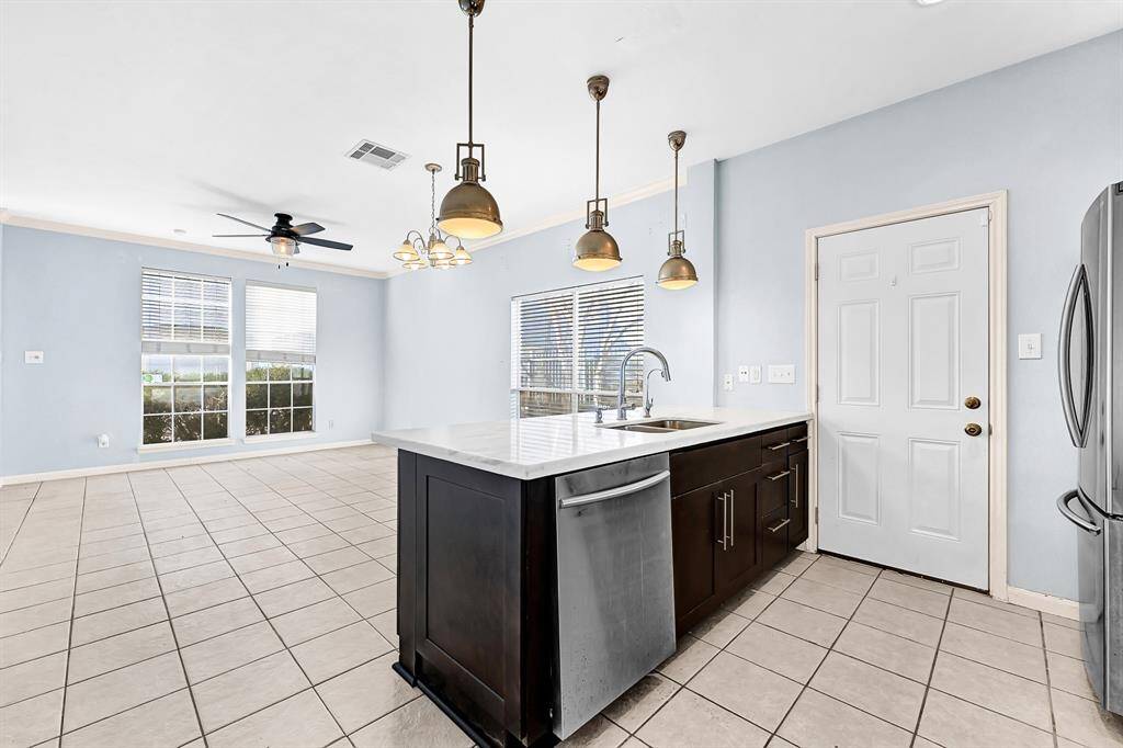 This photo shows a bright, open-plan kitchen and living area. Features include a modern kitchen island with a sink, stainless steel appliances, pendant lighting, and large windows providing ample natural light. The space has light-colored tiled flooring and soft blue walls, creating a welcoming atmosphere.