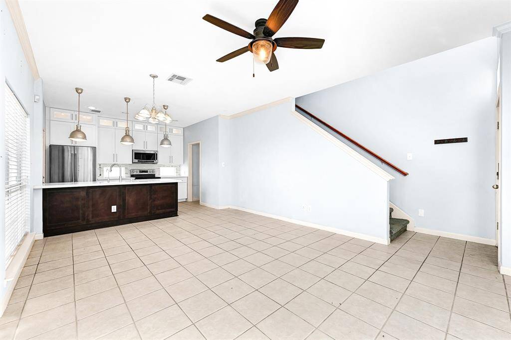 This bright, open-concept living space features a modern kitchen with stainless steel appliances, a central island, and ample cabinetry. The area is tiled throughout and includes a ceiling fan, with a staircase leading to the upper level.