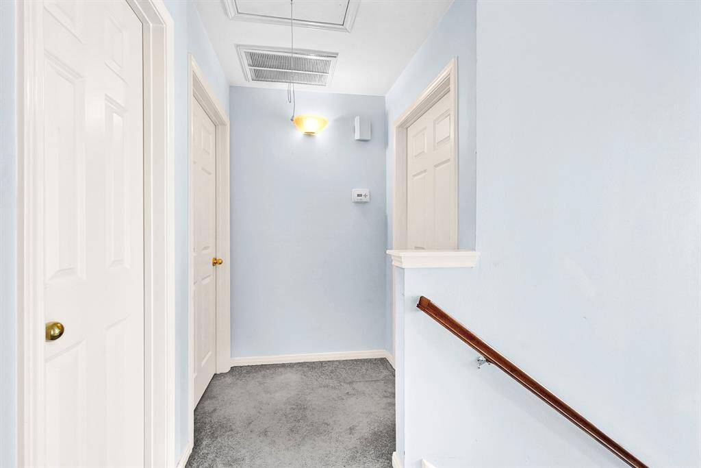 This photo shows a bright hallway with soft blue walls and carpeted flooring. It features multiple doors with brass handles and a wooden handrail leading down the stairs. A ceiling light fixture provides illumination.