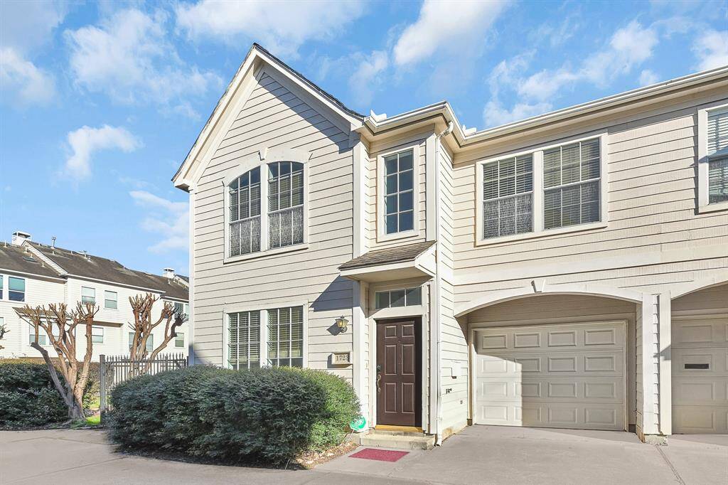 This is a charming two-story townhouse with a light-colored exterior, featuring large windows and a welcoming entryway. It includes an attached garage and is situated in a well-maintained neighborhood.