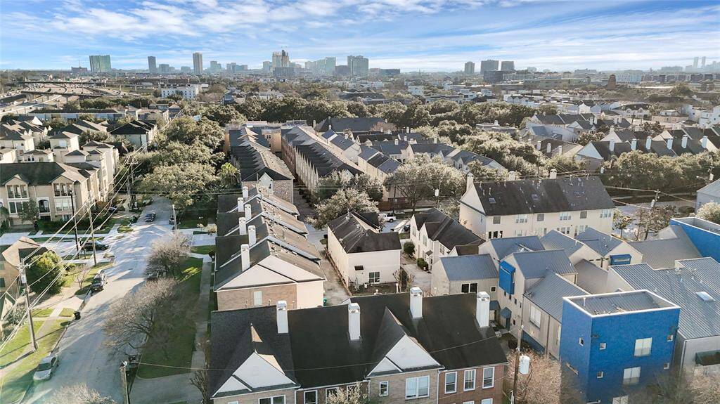 This aerial photo showcases a densely populated residential neighborhood with a mix of townhouses and apartment complexes. The area is lush with trees, providing a suburban feel, while city skyscrapers are visible in the distance, indicating proximity to an urban center. Streets are lined with well-maintained homes, offering a blend of community living and city access.