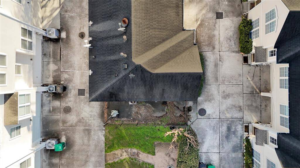 This aerial photo shows a residential area with several houses closely spaced, featuring concrete driveways and small green spaces. The roofs are a mix of dark and light shingles, and the area appears well-maintained with some landscaping visible.