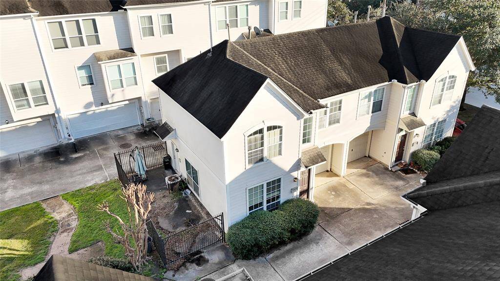 This photo shows a modern two-story townhouse with a shared driveway and attached garage. The exterior is white with large windows, and there is a small fenced yard area on the side. The property is part of a well-maintained complex with greenery and a clean surrounding area.