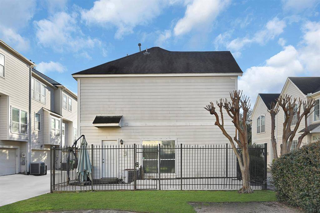 This photo features a modern, multi-story home with a fenced backyard area. The exterior is clad in light-colored siding, and there are well-maintained, pruned trees and a small patio space. The surroundings include similar styled homes, creating a cohesive neighborhood feel.