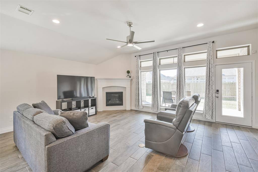 Living room with corner fireplace, wood look tile, LED lights, upgraded ceiling fan, and floor outlet is enhanced by a wall of windows looking out to the covered extended porch.
