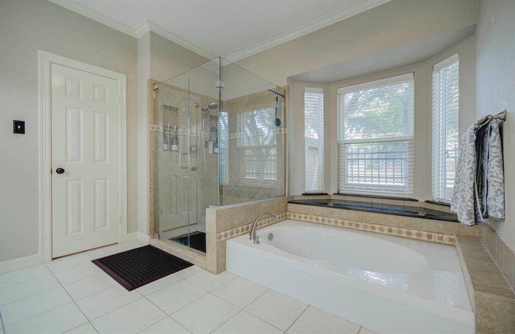 This bathroom features a spacious walk-in glass shower and a luxurious soaking tub set beneath large windows that provide ample natural light. The neutral tile and sleek fixtures create a modern and inviting atmosphere.
