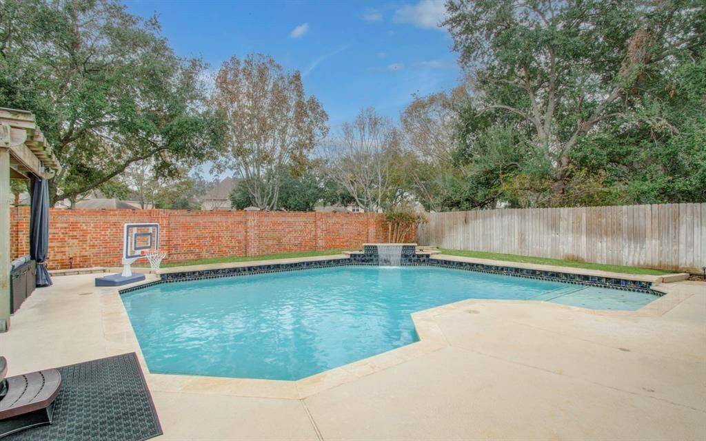 This backyard features a spacious, inviting pool with a waterfall feature, surrounded by a concrete patio and privacy fencing. Lush trees provide a serene atmosphere, perfect for relaxation or entertaining.
