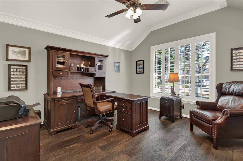 This photo showcases a well-lit home office with a vaulted ceiling, featuring dark wood furniture, including a desk, chair, and bookshelves. Large windows with shutters allow natural light, while the room is accented with framed art and a ceiling fan.