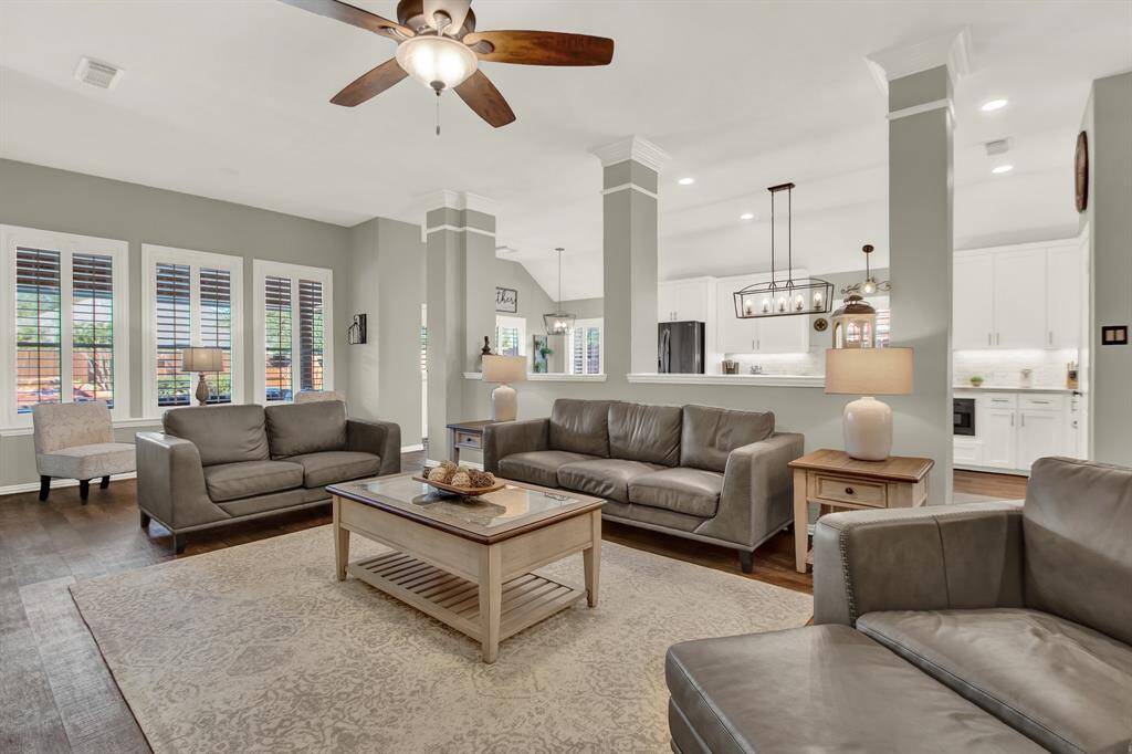 This spacious living area features a cozy seating arrangement with leather sofas and a central coffee table. It offers an open layout with a view into the modern kitchen, highlighting white cabinetry and contemporary lighting. Large windows with shutters provide ample natural light, and the neutral color palette creates a welcoming ambiance.