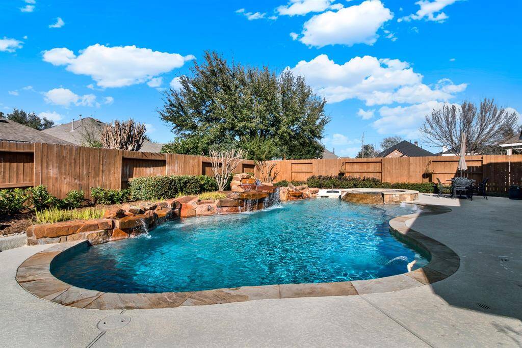 This photo showcases a spacious backyard with a beautiful in-ground swimming pool and rock waterfall surrounded by a wooden privacy fence, and landscaped greenery.
