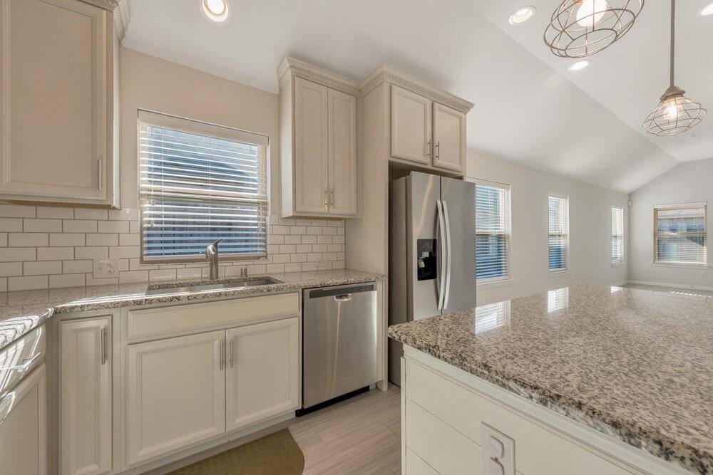 This kitchen features a modern design with granite countertops, stainless steel appliances, and white cabinetry. The open layout and large windows allow for ample natural light, enhancing the spacious feel. Pendant lighting adds a stylish touch.