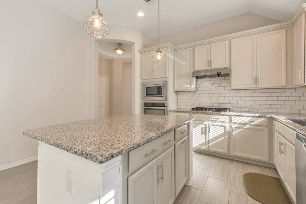 This modern kitchen features granite countertops, white cabinetry, and stainless steel appliances. It has a subway tile backsplash, a central island with pendant lighting, and a built-in microwave and oven. Bright and inviting space.