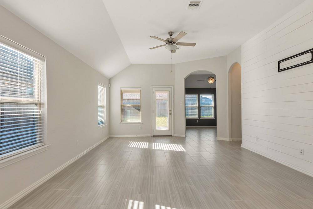 Bright and spacious living area with large windows, modern tile flooring, and a ceiling fan. The room features a neutral color palette and opens to an adjoining space through an archway, leading to a view of the backyard.
