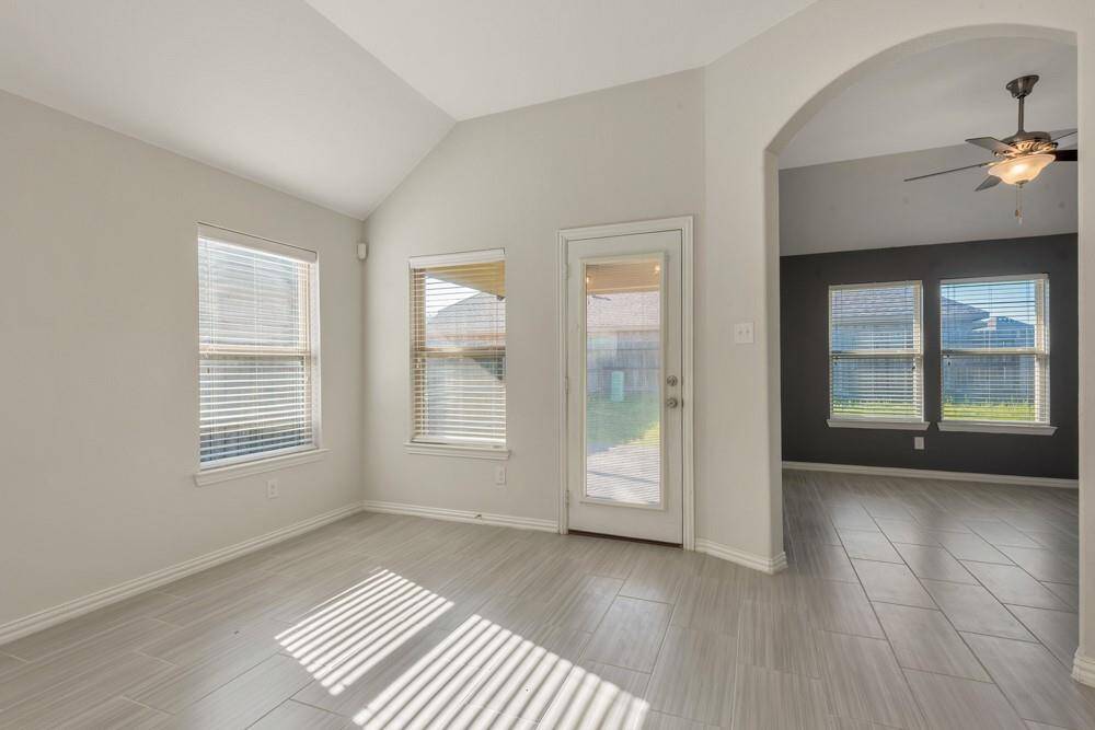 Bright, open room with large windows and modern tile flooring. The space features a door leading to the backyard and an archway connecting to another room with a ceiling fan.