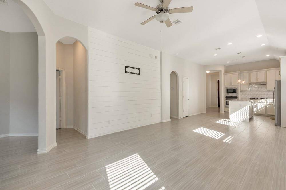 Bright, open-plan living space with high ceilings and modern tile flooring. Features a white shiplap accent wall, ceiling fan, and recessed lighting. The kitchen is equipped with stainless steel appliances and a subway tile backsplash, creating a sleek, contemporary feel.