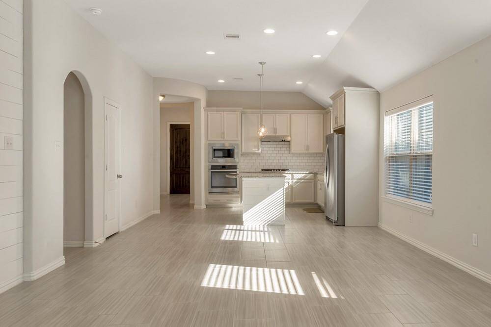 This photo showcases a bright and open kitchen and living area with modern light fixtures, light-colored cabinetry, stainless steel appliances, and ample natural light from large windows. The space features a neutral color palette and a high ceiling, creating an airy and welcoming atmosphere.