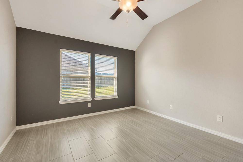 This room features a vaulted ceiling, neutral walls with a dark accent, large windows for natural light, tile flooring, and a ceiling fan. Ideal for a bedroom or home office.