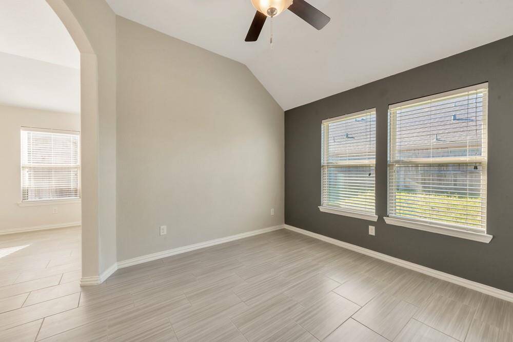 This room features a vaulted ceiling with a ceiling fan, modern tile flooring, and two large windows for natural light. The neutral walls include a dark accent wall, enhancing the space's contemporary feel. An open archway connects to an adjacent area, providing a spacious layout.