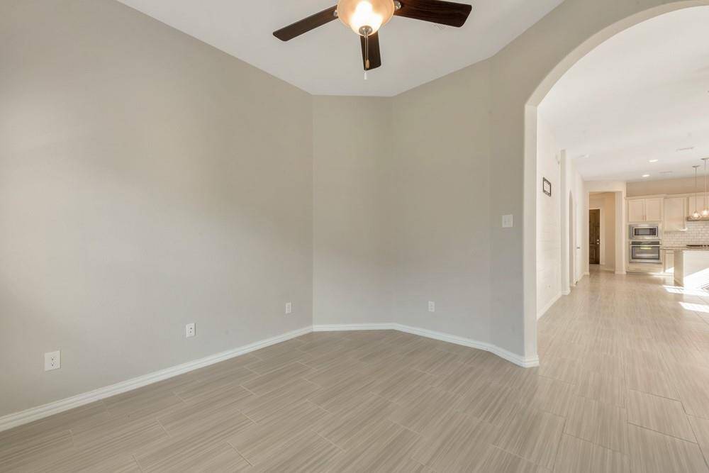 This photo showcases a bright, open space with light-colored tile flooring and neutral walls. It features an arched doorway leading to a modern kitchen area equipped with stainless steel appliances and pendant lighting. A ceiling fan adds comfort to the room.