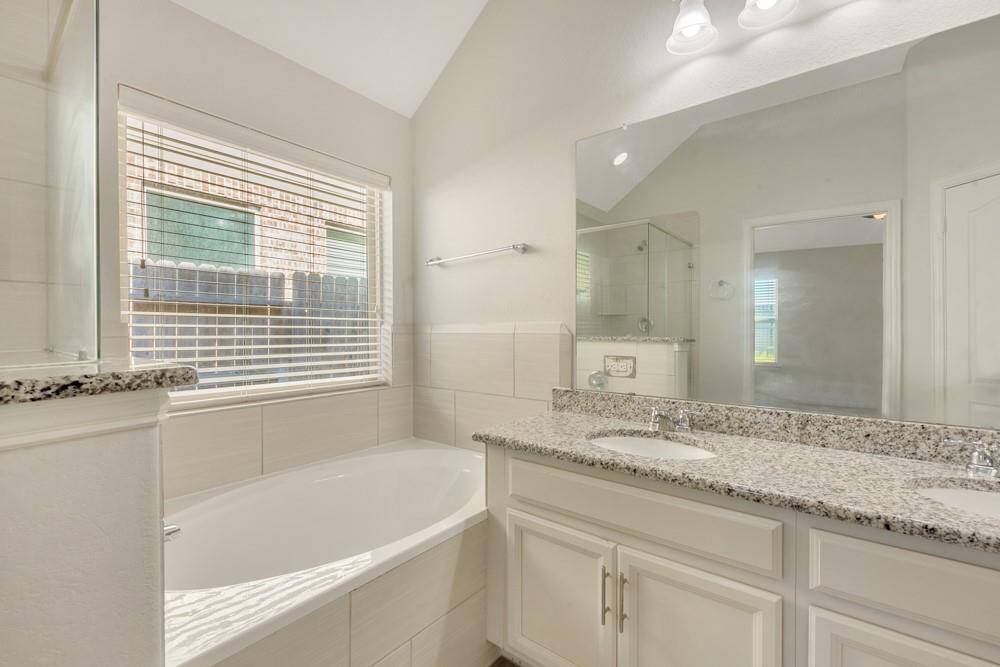 This bright bathroom features a large soaking tub, dual sinks with granite countertops, and a spacious mirror. Natural light enters through a window with blinds, creating a welcoming atmosphere.