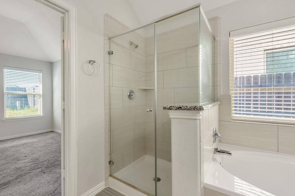 This photo shows a bathroom featuring a glass-enclosed shower with tiled walls, a bathtub with a granite ledge, and a window providing natural light. Adjacent is a doorway leading to a carpeted room with a large window.