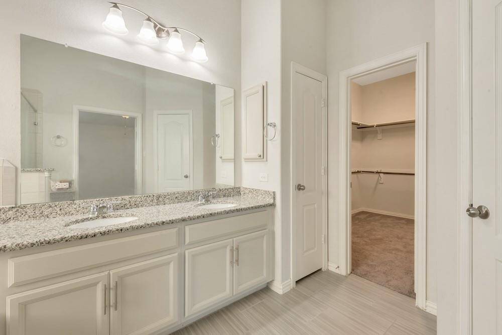 This bathroom features a spacious double vanity with granite countertops, ample under-sink storage, and a large mirror. It includes modern lighting and opens to a sizable walk-in closet, providing excellent storage and convenience.