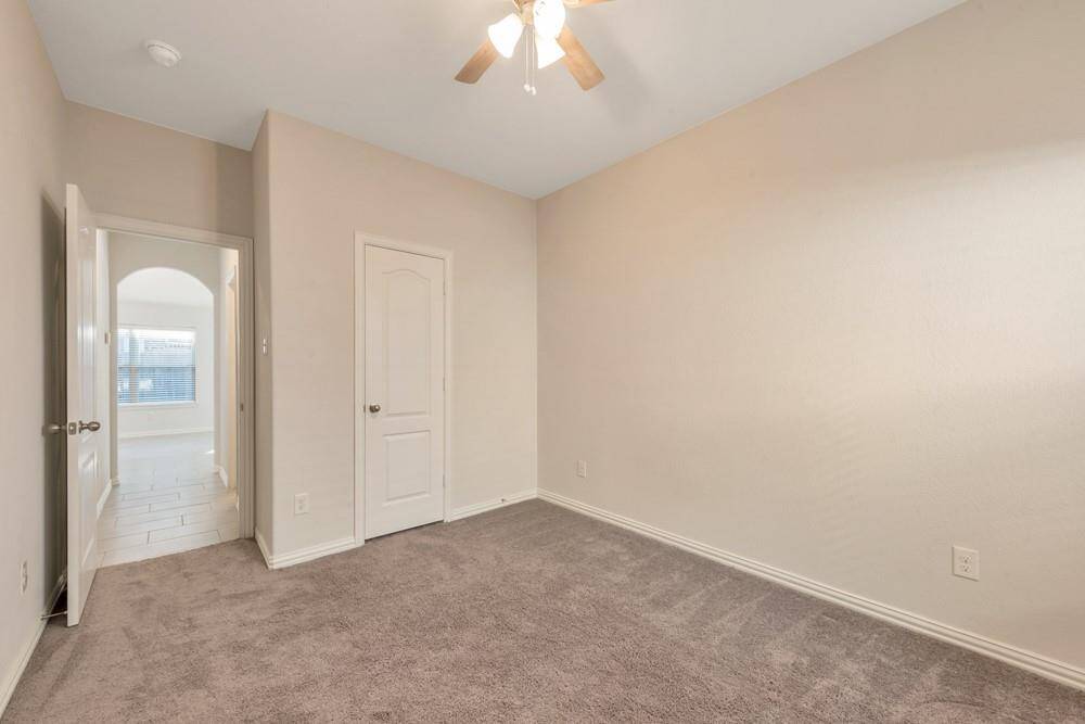 This is a cozy, carpeted bedroom with neutral walls and a ceiling fan. It features a closet with a paneled door and an adjoining hallway with tile flooring leading to a bright, windowed area.