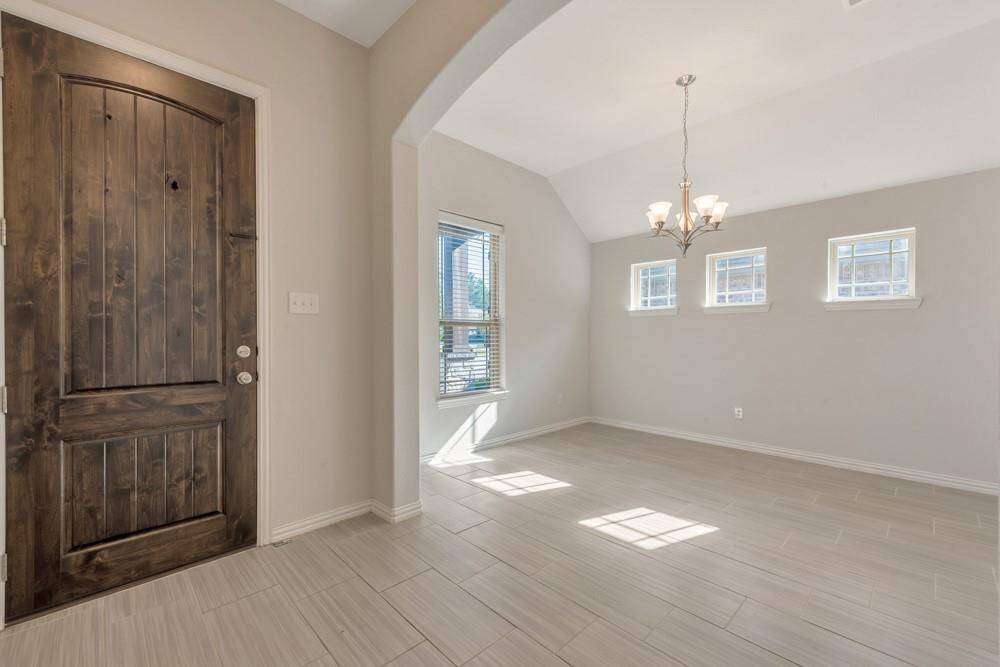 This photo showcases a bright entryway with a rustic wooden door, leading into a spacious room featuring a high ceiling and modern chandelier. The room is well-lit with natural light from multiple windows, and the light-colored tile flooring adds a contemporary touch.