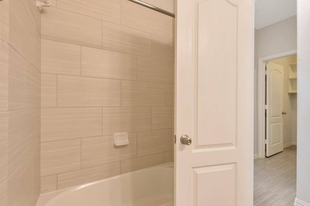 This photo showcases a bathroom with a tiled shower and bathtub, featuring light-colored tiles. The room is accessed through a white paneled door, leading to a hallway with additional doors. The space appears clean and modern.