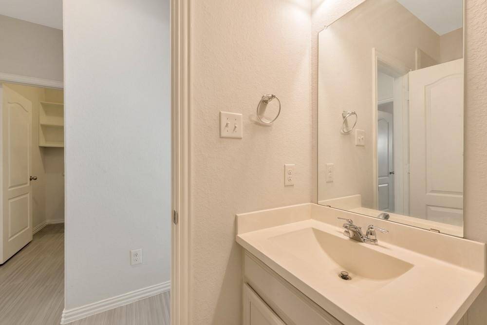 This photo features a bright, modern bathroom with a single vanity, large mirror, and neutral tones. Adjacent to the bathroom is a door leading to a closet with built-in shelving.