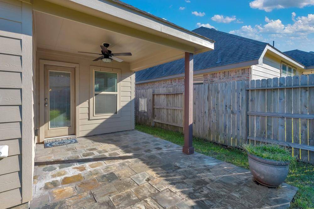 This photo shows a cozy, covered patio area with a ceiling fan and stone flooring. It's enclosed by a wooden fence, offering privacy and a small grassy area, ideal for outdoor relaxation or entertaining.