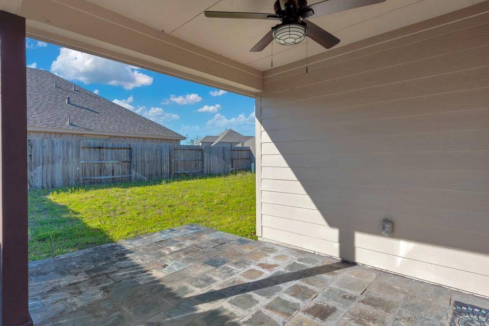 This photo shows a covered backyard patio with a ceiling fan and tile flooring, overlooking a grassy yard with a wooden fence, ideal for outdoor relaxation.