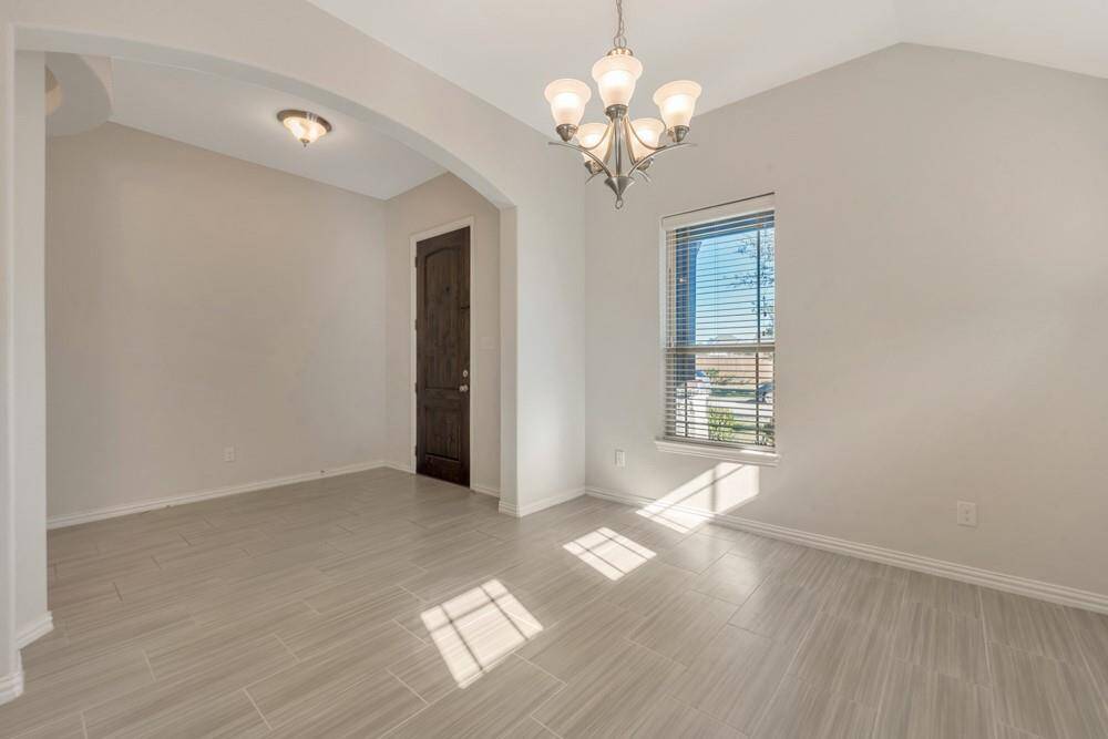 Bright and inviting room featuring large windows, neutral-toned tile flooring, and a stylish chandelier. The arched entryway leads to a dark wooden door, adding a touch of elegance. Ideal for a dining area or living space.