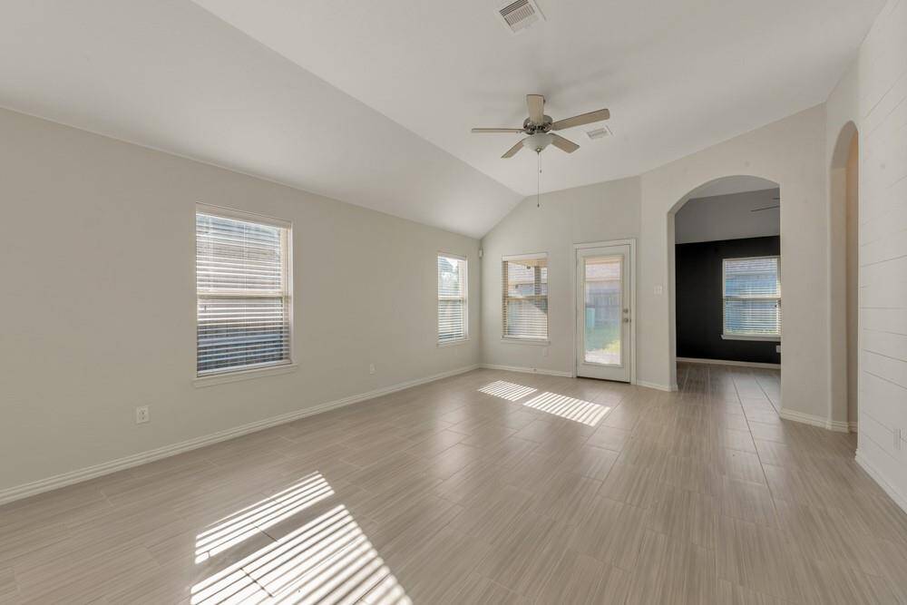 This spacious living area features large windows for ample natural light, neutral-toned walls, and elegant tile flooring. A ceiling fan adds comfort, and the room leads to an adjoining space through an arched doorway.