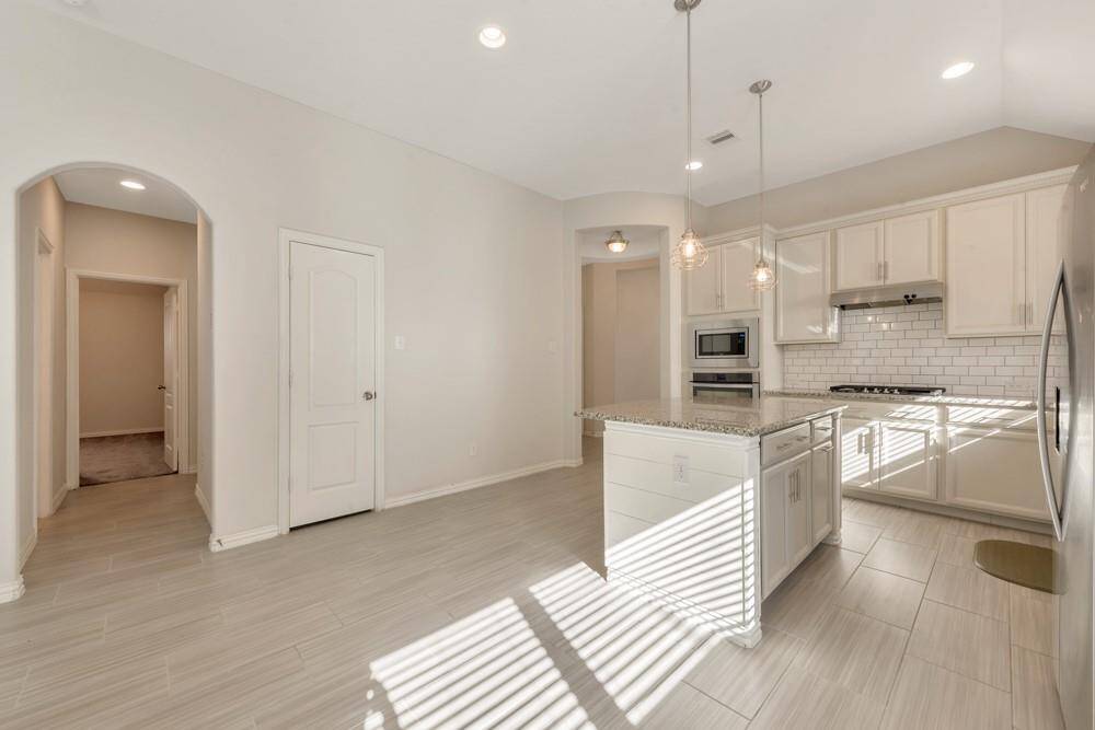 Modern kitchen with sleek white cabinetry and subway tile backsplash. Features an island with granite countertops and pendant lighting. Stainless steel appliances enhance the contemporary feel. The open layout includes tiled flooring and an archway leading to additional rooms.