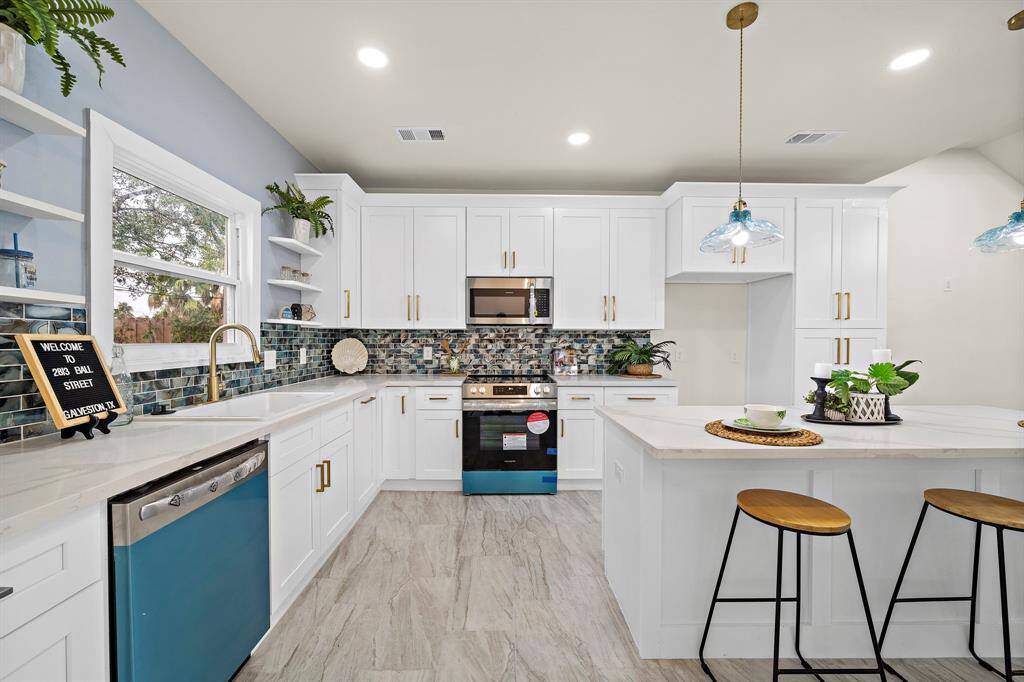 This is a modern, bright kitchen featuring white cabinetry, stainless steel appliances, a stylish backsplash, and an island with bar seating. It has ample natural light and elegant pendant lighting.