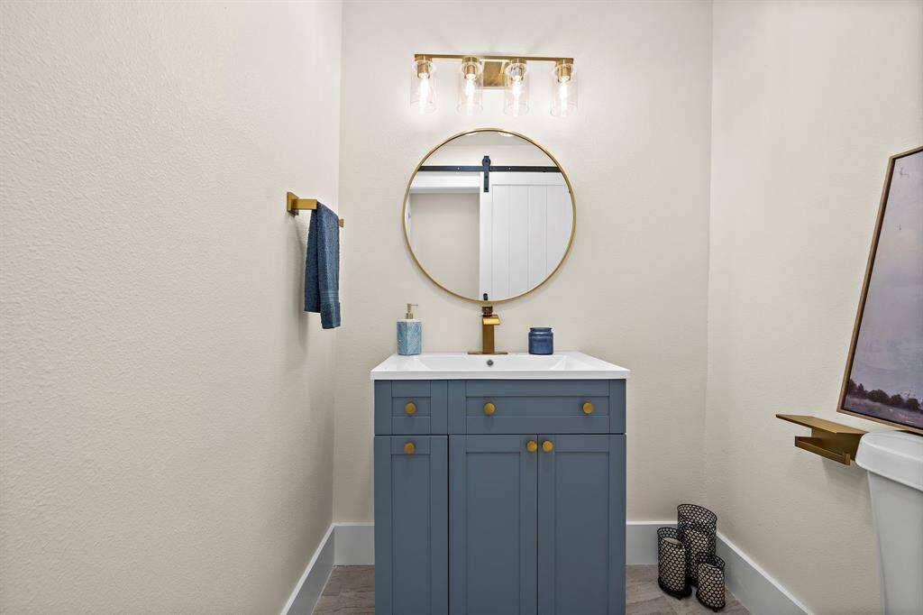 This modern, pristine bathroom features a stylish blue vanity with gold handles, a round mirror with gold trim, and a sleek lighting fixture. It also includes a towel bar and toilet, set against neutral wall colors that provide a serene backdrop.