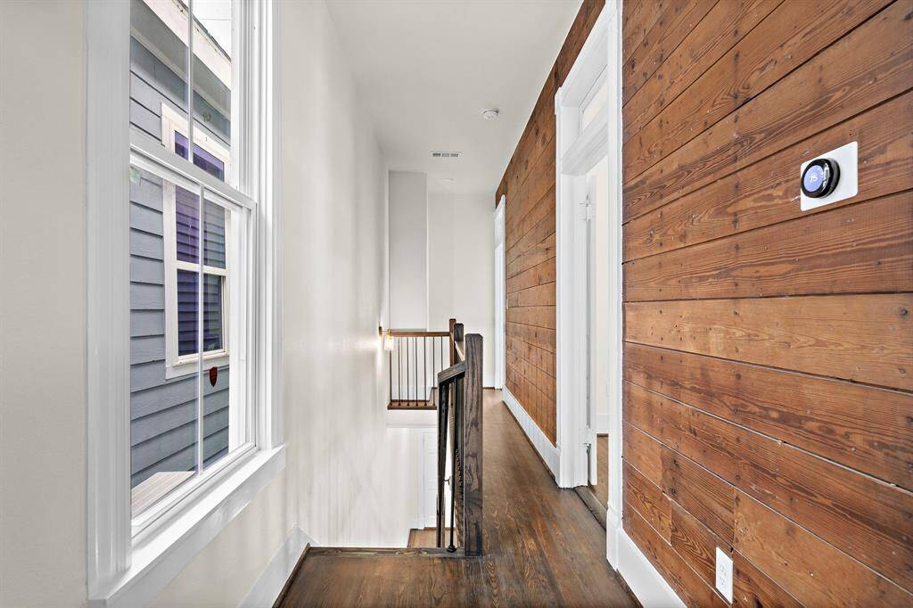 This bright hallway combines modern and rustic elements, featuring natural wood plank walls on one side, white walls on the other, and dark original hardwood floors. A large window allows natural light to flood the space, while a smart thermostat offers convenient climate control.