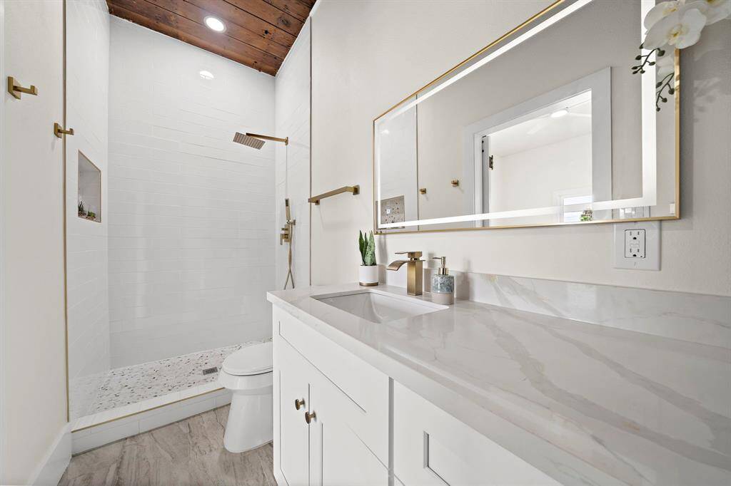 This is a modern bathroom featuring a spacious walk-in shower with gold-tone hardware, and a wooden ceiling beam that adds warmth. The vanity has a quartz countertop and a large mirror with a gold frame, complementing the fixtures. The floor is finished in a gray wood-look tile for a contemporary touch.