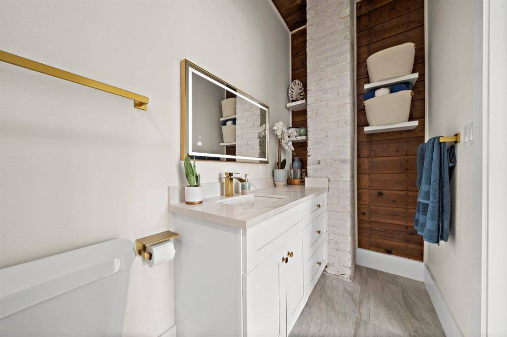 This is a modern bathroom featuring a white vanity with a quartz countertop, brass hardware, a large mirror, and exposed brick accents. There's a built-in shelving nook with wooden back paneling for storage, and the space is complemented by a neutral color palette.