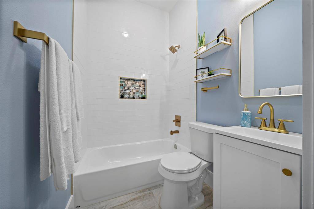 This modern bathroom features crisp white subway tiles, a bathtub/shower combo, a sleek vanity with gold accents, and stylish open shelving for storage.