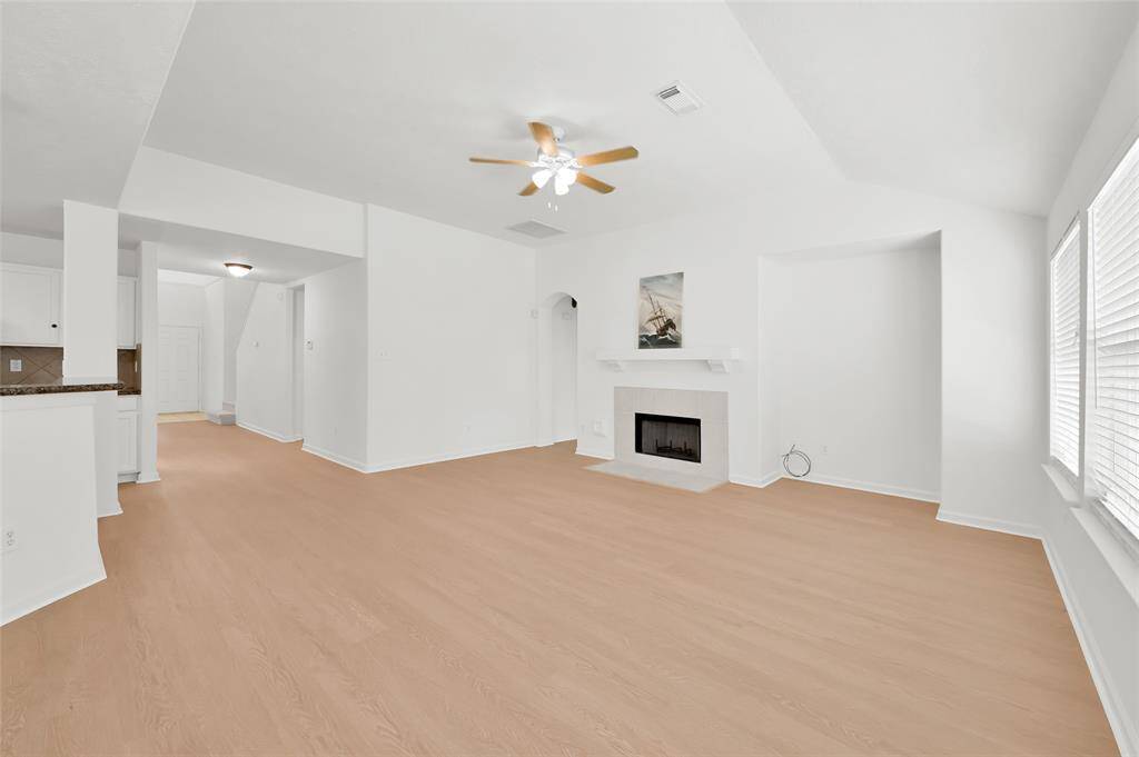 This spacious living area features light wood flooring, a cozy fireplace, and large windows that bring in ample natural light. The open layout flows seamlessly into the adjacent kitchen area, creating an inviting space for family gatherings.
