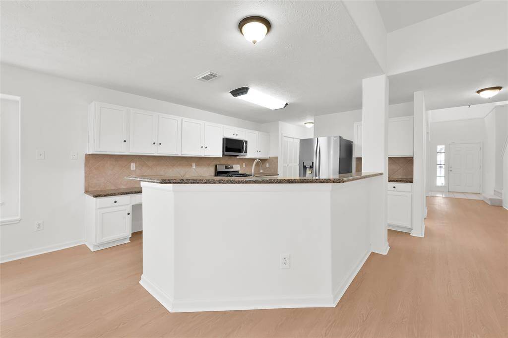 This photo shows a spacious, bright kitchen with white cabinetry, granite countertops, and stainless steel appliances. The open layout leads into the main living area, featuring light wood flooring and ample natural light.