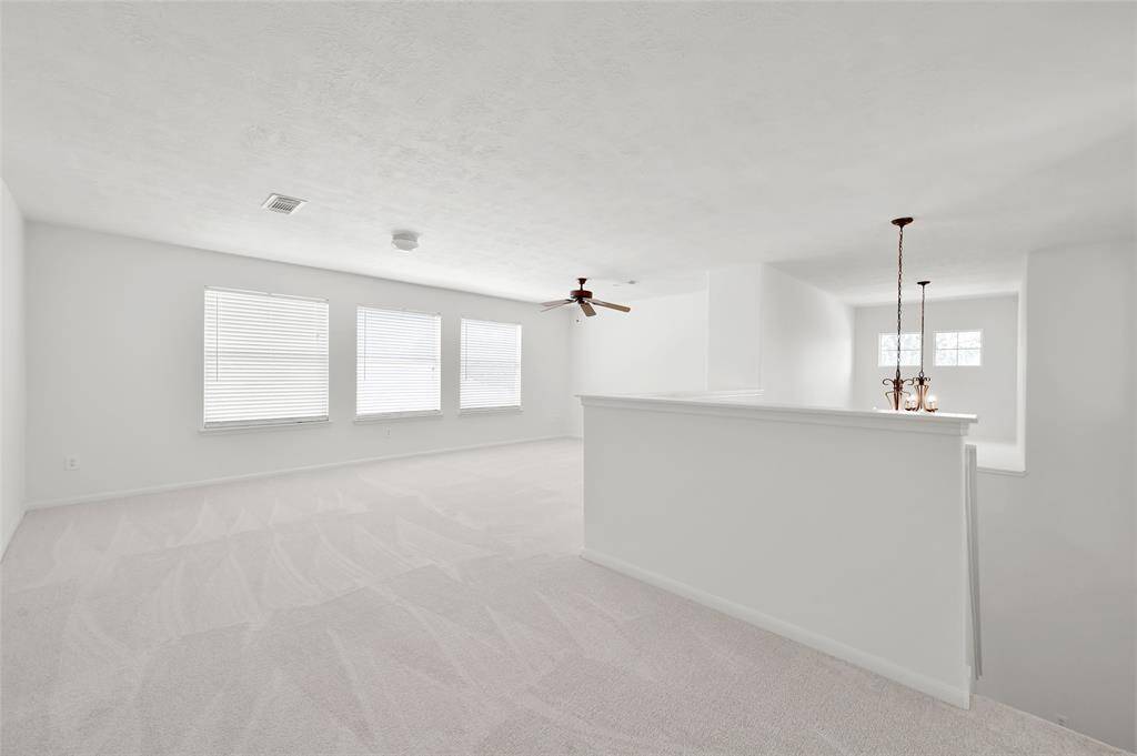 This photo showcases a spacious, bright gameroom with neutral tones, featuring a ceiling fan, multiple windows for natural light, and modern light fixtures. The open layout and white walls create an airy and inviting atmosphere, ideal for personalizing your space.
