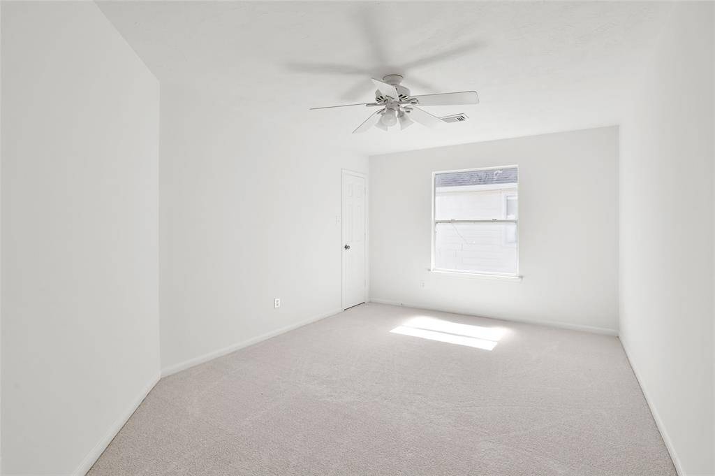 This bright, minimalist room features white walls, a ceiling fan, and soft carpet. A window lets in natural light, and a door provides access to the room.