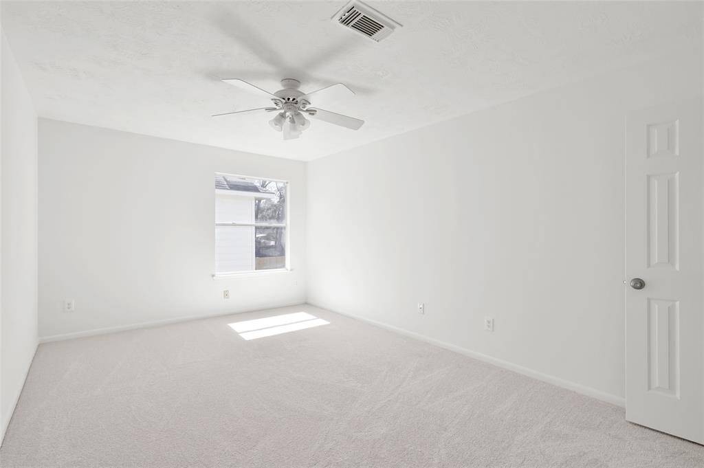This photo shows a bright, empty room with white walls and carpet, featuring a ceiling fan and a window allowing natural light. Ideal for customization.